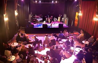 People sitting at individual tables in front of the stage at the Chapel Arts Centre in Bath