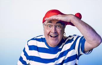 Danny Baker in a blue and white striped t-shirt and a red hat. He is holding his hand to his forehead.