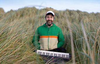 David O'Doherty sitting in tall grass with a small keyboard. Text reads "David O'Doherty, Tiny Piano Man"