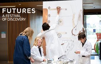 A group of children wearing lab coats, along with a researcher facilitator are crafting a display of urban planning using household items.