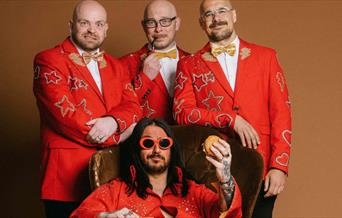 A photo of the band Elvana, they are clad in red, one of the singers in sitting on a chair holding a burger.