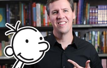 Author Jeff Kinney stands in front of a bookshelf, with a cut-out of the character Greg Heffley in front of him.