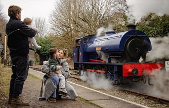 A family of four can be seen from behind waving at a blue steam train from the station platform.