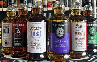 6 bottles of whisky sitting on a countertop with a wall  of spirits behind the,. The whiskies are Springbank 10 Amontillado 55%, Springbank 15 46%, Sp