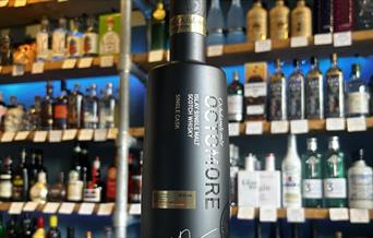 An image of a sleek black bottle of Octomore whisky sitting on a countertop with a colourful wall of spirits behind it.