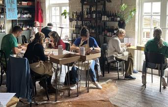 a seated group of people crafting at various tables in a spacious room lined with shelves of wine