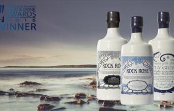 An image of a misty rocky beach off the coast of scotland. The image has 3 bottles of rock rose gin on it. Ceramic white bottles with blue writingg