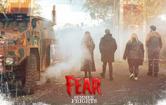 Visitors walking through a foggy, eerie entrance at Fear Summer Frights, featuring an old, rusted military vehicle and industrial structures in the ba