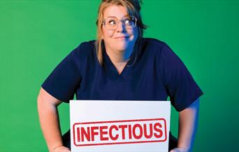 A woman wearing dark hospital scrubs holding a sign that says "infectious" against a green background