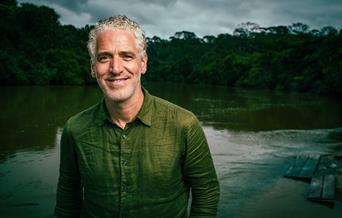 Portrait image of Gordon dressed in a khaki coloured long sleeve shirt, pictured in front of a lake landscape