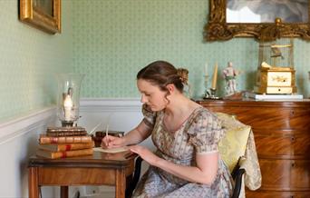 A woman in Regency clothing writing at a writing desk