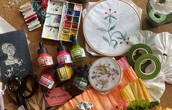 A colourful chaos of crafting tools and materials with a Regency theme sprawled out including lino tools, scissors, threads, embroidery hoops, paints