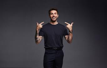 Joel Dommett in a black T-shirt pointing at his face with both hands