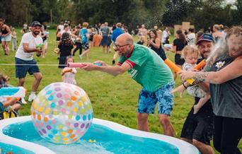 A group of people, including adults and children, are having a lively water fight outdoors on a grassy field.
