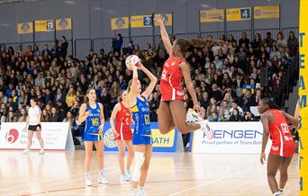A defender, wearing red, leaps into the air as a Team Bath Netball shooter, wearing blue, lines up a shot. There is a stand of spectators watching beh