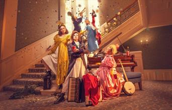 A group of women dressed in period costume. Three are at the bottom of a staircase grouped around a piano, while the other two are on the staircase, w