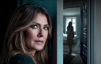 A woman stands behind a door, with a mysterious woman in a white dress in the background, not facing the camera.