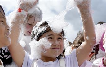 Children playing joyfully in a cloud of foam bubbles under a bright sky. A young girl in the center, smiling with arms raised, is covered in bubbles.