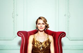 A woman in 1920s attire is sat down on a red chair, she's smiling.