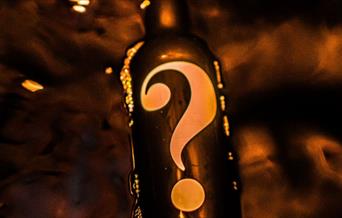 A black whisky bottle with a question mark on it. The background is also dark with smoke and embers of fire floating around the bottle.