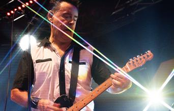 A guitarist plays on stage surrounded by bright stage lights
