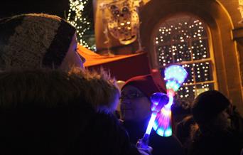 A small boy, wearing a knitted hat and warm furry coat, is holding a multicoloured light up toy. He is in a crowd of people, who are standing in front