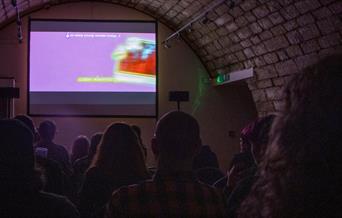 A purple-lit image of a groups of people facing a large screen, showing T-Blockers, a film directed by Alice Maio Mackay.