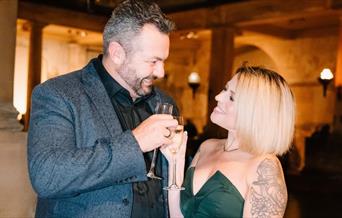 A smiling couple toast each other with a glass of Champagne around the Roman Baths