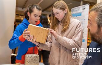 Two young girls study some innovative building materials while a University of Bath researcher explains them