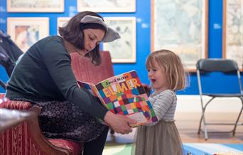 A lady reading wearing a headband with furry ears, reading a book to a young girl.