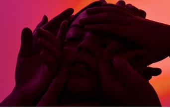 Close-up of a person's face surrounded by several hands, with dramatic red-pink lighting creating a moody, intense feel.