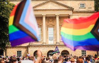 A photograph of the front of the Holburne Museum with a crowd of people in front. Two large progress rainbow flags fly in the foreground.

