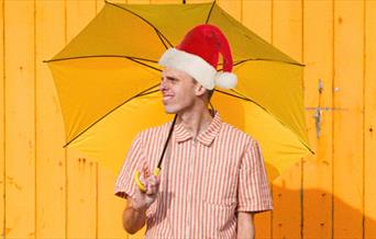 A man dressed in bright colours and a Santa hat holding a yellow umbrella