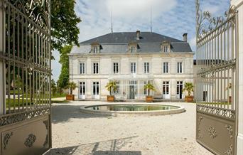 An image of the gated front to a 18th century french cognac chateau. The building is a large stone 3 floor chateau with a circular pool in front and a