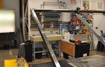 A screen printing studio with various screens, paint containers, and equipment. The setup includes a sink, storage shelves with supplies, and a large