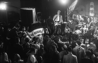 A black and white photo of a band playing live. A person in the crowed is holding up a banner with the words "The Rogues" written on it.