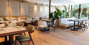 Restaurant with pine floor, white and wood furniture, plants and a pale stone wall. It has a glass fronted side that looks out over a garden.