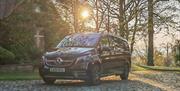 A black Mercedes luxury minivan parked on a cobbled road behind an ivy-covered manor