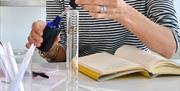 The midsection of a person taking a pipette out of a fragrance bottle, with a measuring cylinder and notebook on the table in front of them.