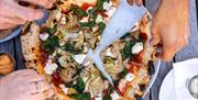 Three pairs of hands tearing off slices of pizza from a plate on an outdoor table