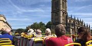 A group of tourists on the top floor of an open-top bus alongside Bath Abbey.