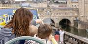 View from behind of a woman and two children sat on an open-top bus, looking and pointing at the surrounding area.