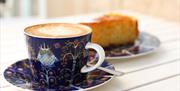 A coffee in a decorative mug, with a slice of cake out of focus behind.