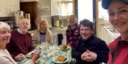 A group people sat at a table eating food with a women in a pink jacket taking their picture