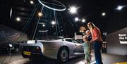 A mum and two kids looking at a silver vintage Jaguar on a podium