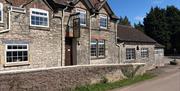 Outside of a stone brick country pub next to a road. It has a dark tiled pitch roof and many small windows. Trees and blue skies are in the background