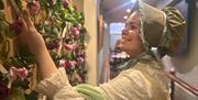 A woman in a regency-era dress and bonnet reaches up towards a flower-covered wall inside the Jane Austen Centre exhibition.