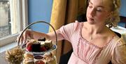 A woman in a pink regency dress reaching out towards a cake on a tiered afternoon tea platter, sat beside a window in a dining room with blue walls.