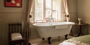 A claw-foot bath tub next to a large window with white curtains.