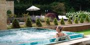 Lady in an outdoor pool looking over a manicured garden with small topiary bushes, a white parasol and sunbeds.